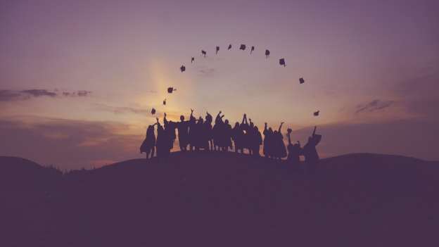 The silhouette of a group of people during sunset on a hill throwing their graduation hats into the air. The hats form an arch above the group of people.